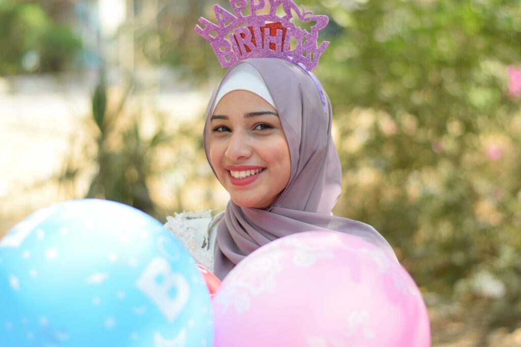 Smiling woman wearing hijab and birthday hat, holding colorful balloons outdoors, celebrating a joyful birthday.