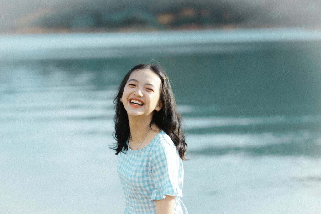 A cheerful young woman in a plaid dress laughing by a serene lake.