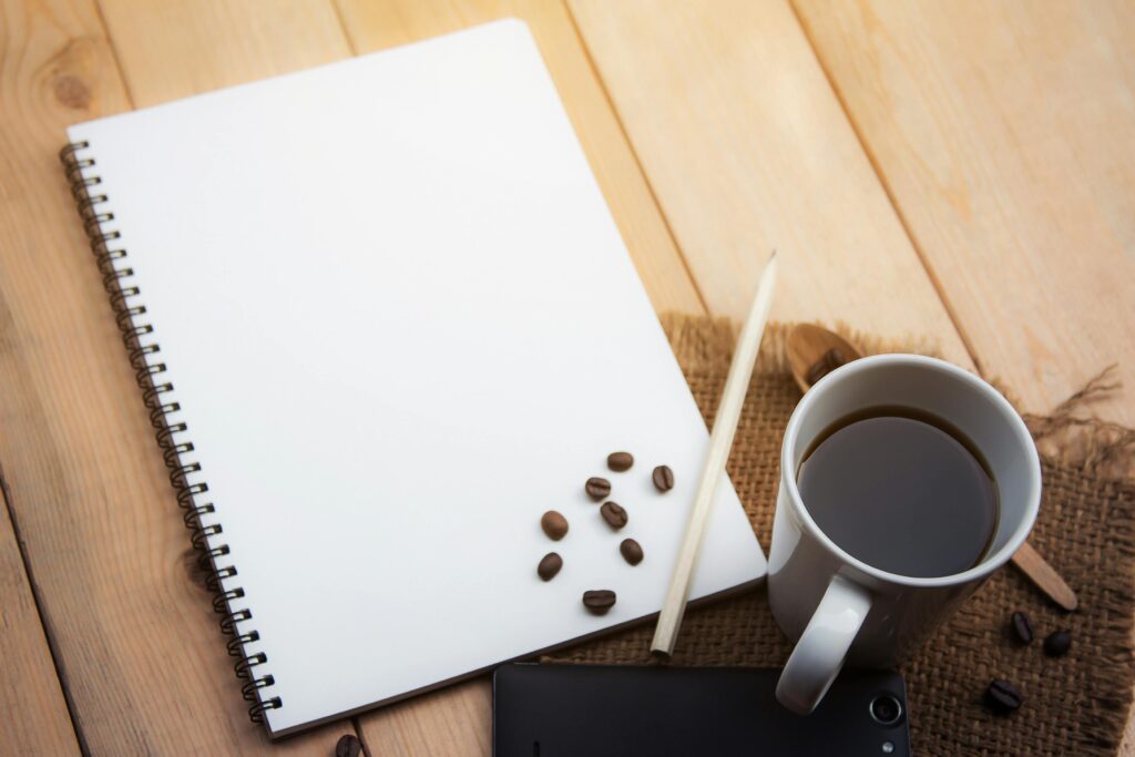 Spiral notebook, coffee cup, pencil, and smartphone on wooden desk, ideal for office or creative work concepts.