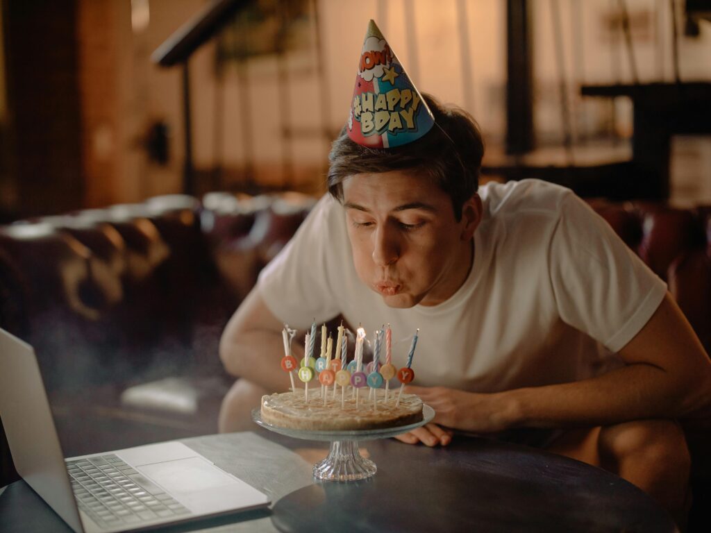 A young man celebrates his birthday with a cake and a laptop in a cozy home setting.